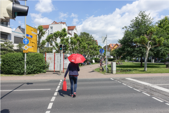 MTV: Ihr Weg zu uns! Eine Person mit einem roten Regenschirm und einer roten Tasche überquert bei Grünlicht einen Zebrastreifen. Im Hintergrund sind Wegweiser, Bäume und ein kleiner Park zu sehen.