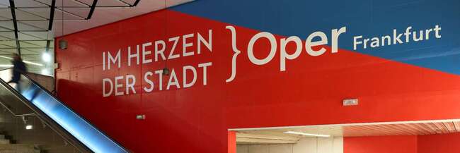 Eine Rolltreppe, rot-blaue Wände, Schrift: Im Herzen der Stadt Oper Frankfurt