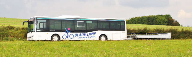 Vergrößerte Ansicht: Ein weißer Bus der Blauen Linie mit Fahrradanhänger fährt durch eine sommerliche Landschaft