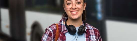 Mädchen mit dunkelrot-kariertem Hemd und Kopfhörern um den Hals vor einem Bus