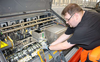 Junger Mann gebückt vor einem großen grauen Kasten mit vielen Kabeln und Drähten. Zwei gelbe Warnschilder mit Blitz-Symbol. Davor ein kleines gelbes Messgerät.