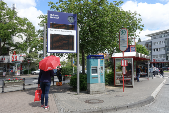 MTV: Ihr Weg zu uns! Eine Bushaltestelle mit einer elektronischen Anzeigetafel für kommende Busverbindungen am „Hofheim Bahnhof“. Eine Person mit einem roten Regenschirm und einer roten Tasche wartet unter der Anzeigetafel. Rechts neben ihr befindet sich ein Fahrkartenautomat. Im Hintergrund sind Geschäfte und Straßenverkehr zu sehen.