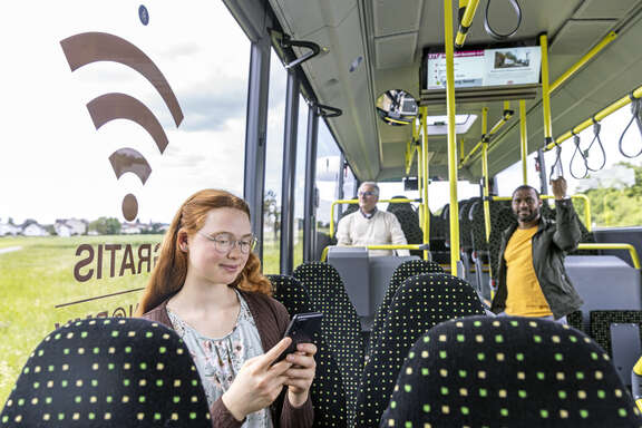 Mädchen mit Smartphone sitzt in einem Bus. Auf der Fensterscheibe klebt das WLAN-Symbol.