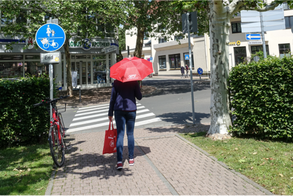 MTV: Ihr Weg zu uns! Eine Person mit einem roten Regenschirm und einer roten Tasche geht auf einem gepflasterten Gehweg in Richtung eines Zebrastreifens. Ein blaues Schild zeigt das Ende eines kombinierten Geh- und Radwegs an. Links steht ein Fahrrad, im Hintergrund befindet sich eine Apotheke und ein Fußgängerüberweg. Der Bereich ist von grünen Bäumen und Hecken umgeben.