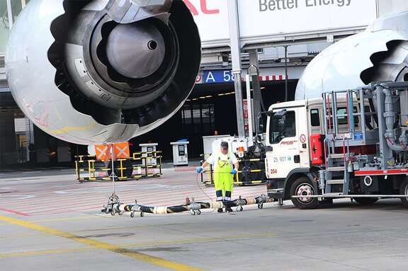 Vergrößerte Ansicht: Mann arbeitet an Tankschlauch neben Lastwagen, über ihm zwei riesige Turbinen