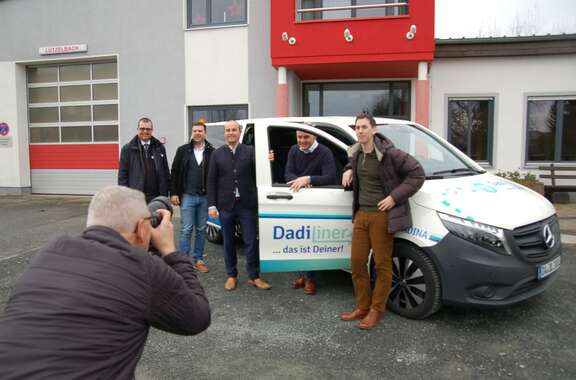 Fünf Männer stehen an einem weißen Auto mit der Aufschrift: DadiLiner. Im Vordergrund macht ein Mann ein Foto davon.