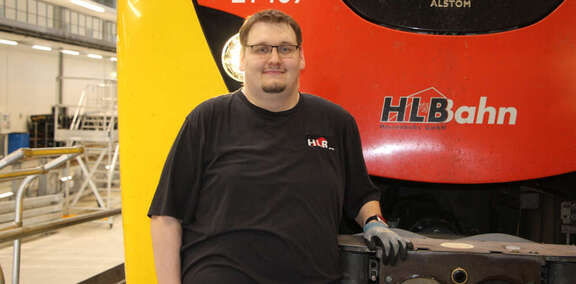 Junger Mann mit Brille steht vor einem schwarz-rot-gelben Zug. Neben ihm die Aufschrift "HLB Bahn"