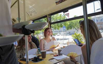 Drei junge Frauen sitzen mit Fächer in der Tram, links eine Person mit drei Tellern in der Hand