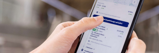 Smartphone von zwei Händen gehalten. Auf dem Bildschirm ist eine Fahrplanauskunft zu sehen.