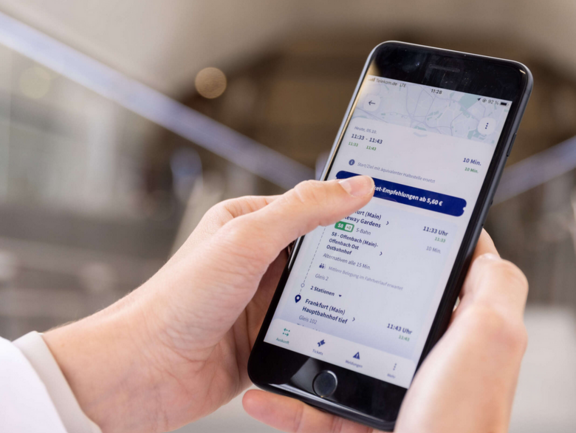 Smartphone von zwei Händen gehalten. Auf dem Bildschirm ist eine Fahrplanauskunft zu sehen.