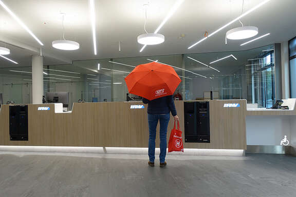 MTV: Ihr Weg zu uns! Die Person mit dem roten Regenschirm steht in einem modernen, hell beleuchteten Empfangsbereich mit einer langen Holztheke. Auf der Theke befinden sich Logos von 'RMV'.