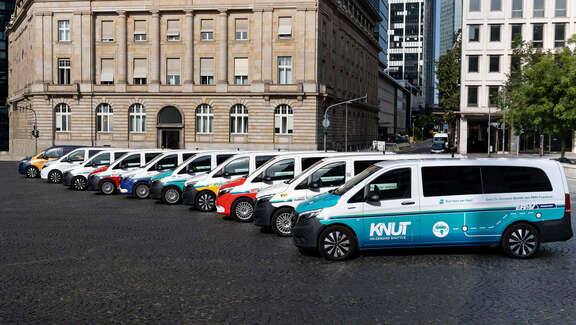 Verschiedene Fahrzeuge stehen in einer Reihe versetzt vor einem Gebäude. Seitliche Sicht auf die Fahrzeugflotte