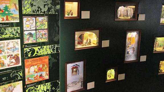 Brüder Grimm-Haus Raum Märchenwald Vergrößerte Ansicht: kleinen Schaukästen an dunkler Wand mit Märchenszenen, links daneben bunte Märchenbilder