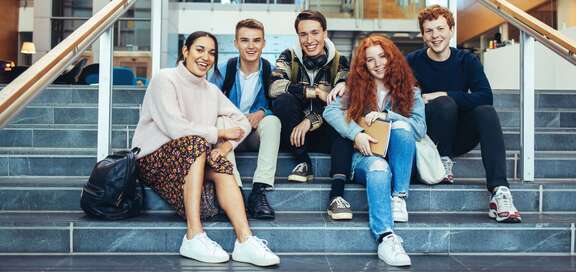 Fünf Schülerinnen und Schüler sitzen in einem Gebäude auf Treppenstufen 