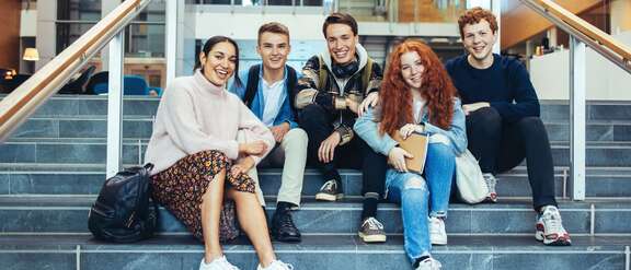 Fünf Schülerinnen und Schüler sitzen in einem Gebäude auf Treppenstufen 