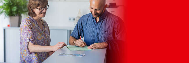 Eine Frau mit Brille und bunter Bluse steht lächelnd an einem Schalter und erklärt einem Mann ein Formular, das er gerade ausfüllt.