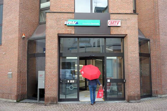 MTV: Ihr Weg zu uns! Die Person mit dem roten Regenschirm steht vor einem Gebäude mit der Aufschrift 'RMV MobilitätsZentrale' und 'MTV'. Der Eingang ist aus Glas und befindet sich in einer Backsteinfassade.