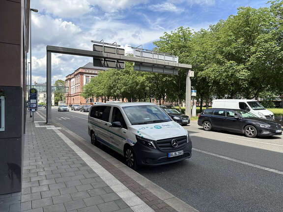 Ein größeres weißes Auto steht auf einer Straße, im Hintergrund Bäume und ein historisches Gebäude