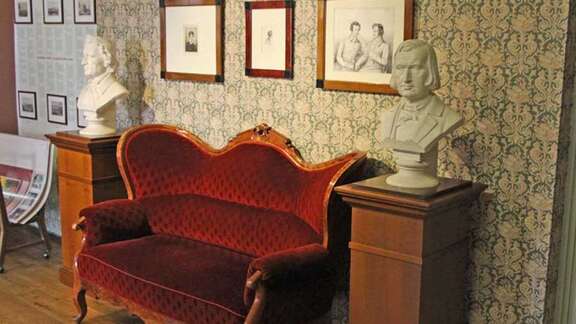 Brüder Grimm-Haus Sofa Eingangsbereich Vergrößerte Ansicht: altes geschwungenes Sofa mit rotem Samtbezug und Holzgestell, flankiert von zwei weißen Büsten