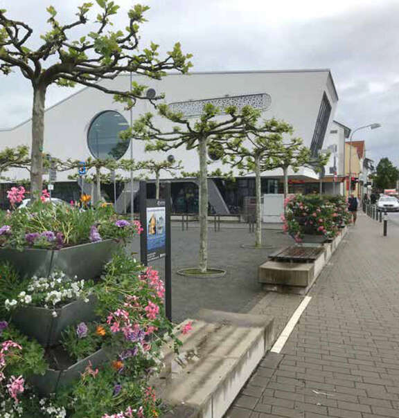 Platz mit Sitzgelegenheiten, Blumen und Bäumen, ein Gebäude im Hintergrund.