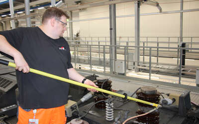 Junger Mann mit orangener Hose und schwarzem Shirt hat eine lange gelbe Stange in der Hand befestigt ein Gerät an einer schwarzen Befestigung