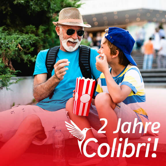 Teaserbild Colibri 2 Jahre "2 jähre Colibri!": Älterer Mann mit Hut und Sonnenbrille sitzt neben einem Jungen mit Kappe. Beide essen Popcorn.
