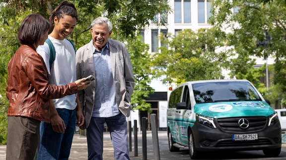 Eine junge Frau zeigt einem jungen Mann und einem älteren Mann ihr Handy. Rechts im Hintergrund steht ein On-Demand-Fahrzeug