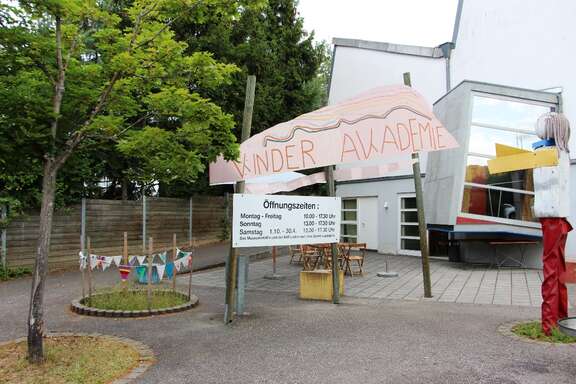 Vergrößerte Ansicht: asymmetrisches Gebäude, bemaltes Schild mit Aufschrift Kinder Akademie, Schild mit Öffnungszeiten