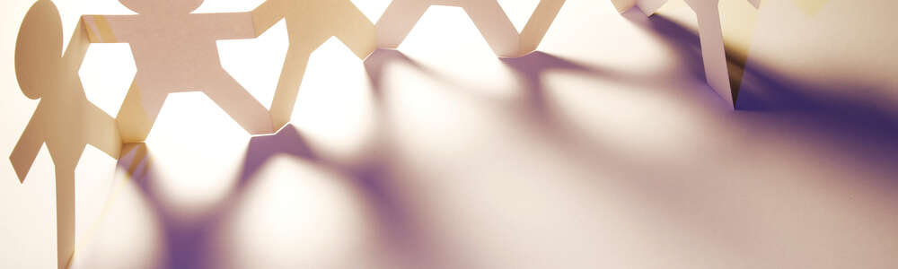 Eine beige Kette Papiermännchen steht auf einem hellen Hintergrund und wirft dunkle Schatten in den Vordergrund