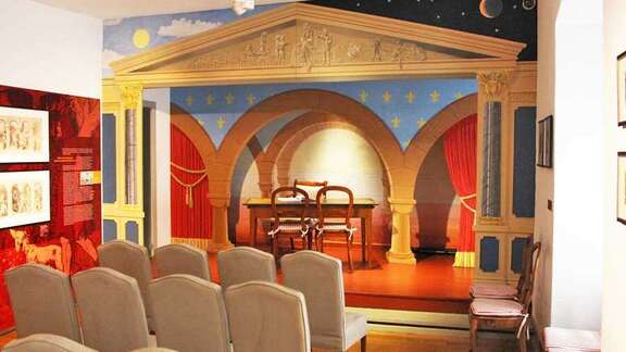 Brüder Grimm-Haus Alter Gerichtssaal Vergrößerte Ansicht: Stuhlreihen vor einer Bühne mit Märchenmotiv