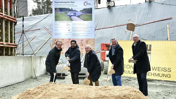 Symbolischer Spatenstich im Sand auf einer Baustelle mit fünf Männern