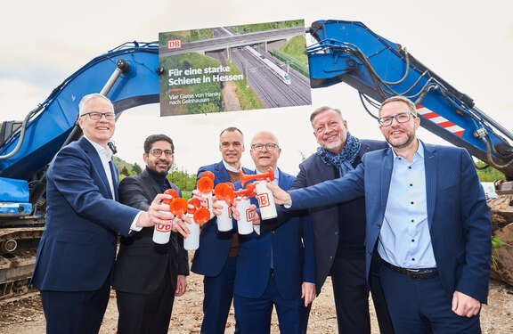 Start viergleisiger Ausbau der Strecke Hanau - Gelnhausen Hinten zwei blaue Bagger, vor denen ein Plakat mit der Aufschrift "Für eine starke Schiene in Hessen. Vier Gleise von Hanau nach Gelnhausen. Im Vordergrund sechs Männer mit Tröten in Hand, auf denen das Logo der Deutschen Bahn zu sehen ist.