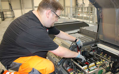 Junger Mann hockt mit Handschuhen neben einem geöffneten grauen Kasten und arbeitet an elektrischen Geräten